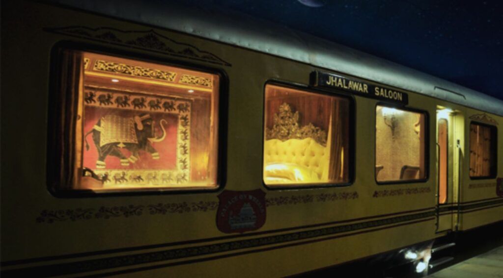   Night view of the Palace on Wheels' Jhalawar Saloon, showcasing the luxurious interior with royal décor, including an elephant mural and opulent furnishings inside a brightly lit yellow train.