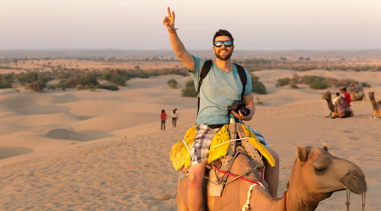 A smiling traveler wearing sunglasses, holding a camera, and sitting atop a camel in the golden sand dunes of India during sunset.