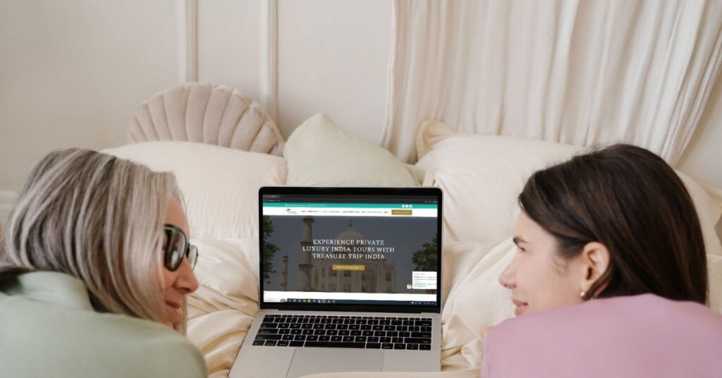 Two friends lying on a bed, looking at a laptop displaying the Treasure Trip India website offering luxury private tours.