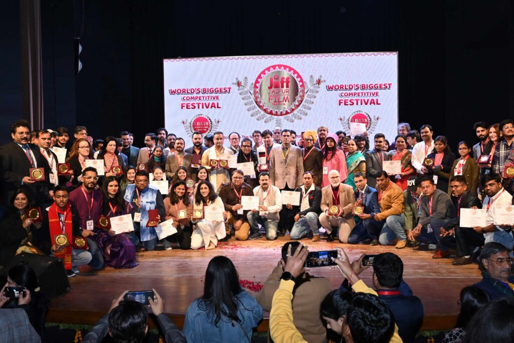 : A group photo of awardees holding certificates and medals on a stage, taken at the Jaipur Film Festival. A large banner in the background reads, "World's Biggest Competitive Festival," highlighting the event's grandeur.