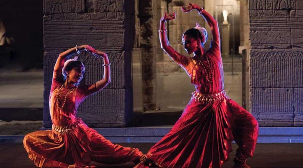 Two women in traditional Indian attire performing a classical dance against a historical backdrop.