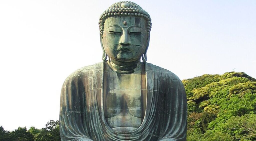  A large, serene Buddha statue made of bronze, with lush green trees and a clear blue sky in the background.