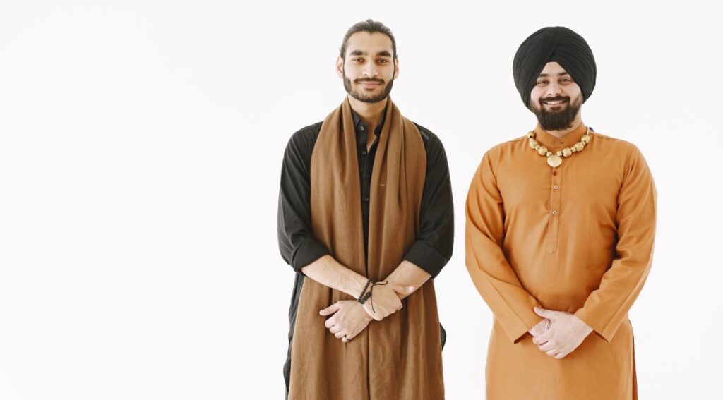 Two men in traditional Indian attire, one wearing a turban and an orange kurta with a necklace, the other in black clothing with a brown scarf.